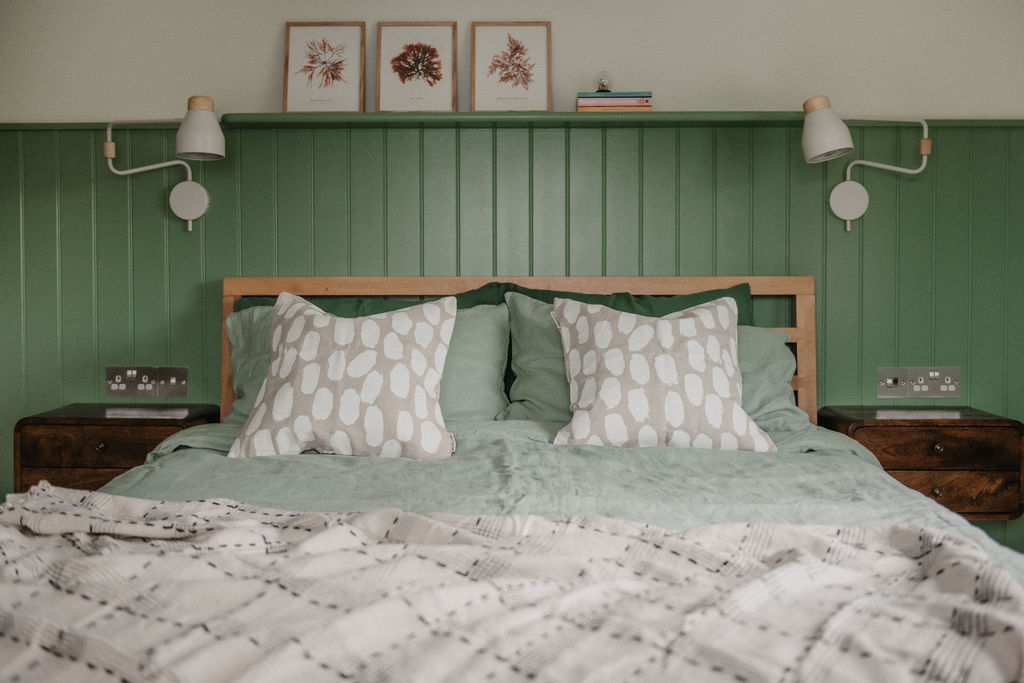 Head of the king bed with 2 scatter cushions and matching bedside lamps. Shelf acroos top of the panelling with 3 picture frames and books.