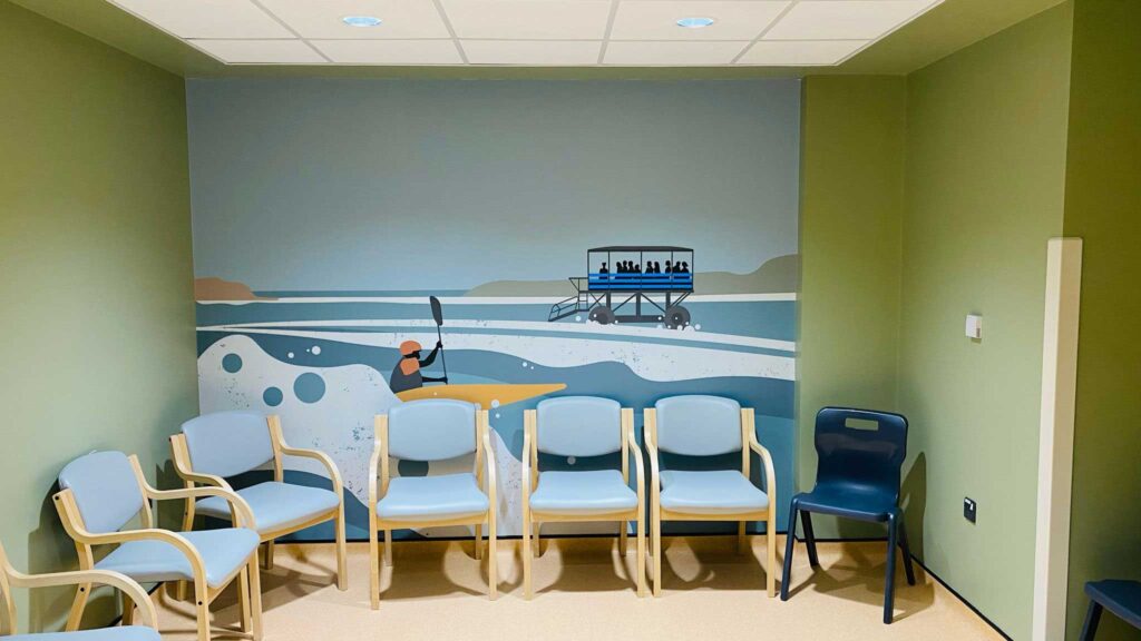 A mural in the waiting room with blue chairs in front. The Sea Tractor travels through the sea to Burgh Island taking passengers. There's a yellow kayaker in the foreground with the waves splashing.