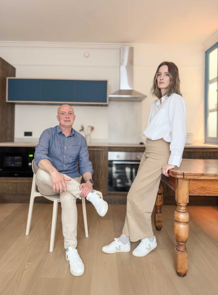 Stephen is seated and Alice stands against a vintage dining table. A kitchen is in the background.