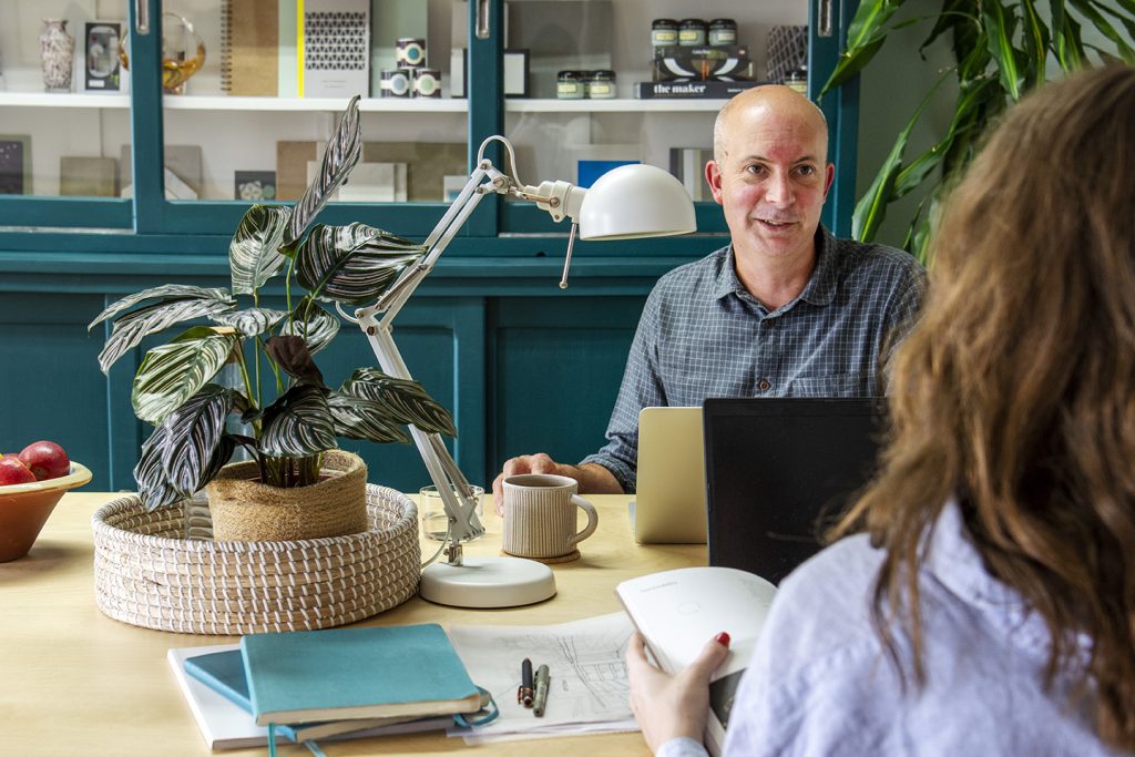 Two workers at a desk in a design studio with a potted plant and a large plant behind them.