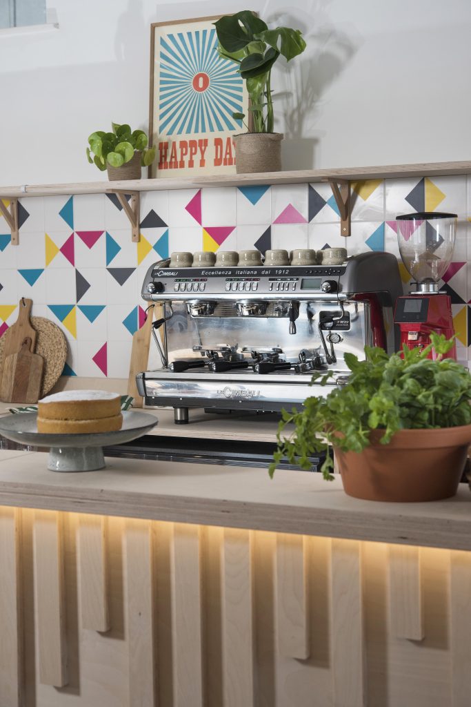 Coffee machine against a colourful tile with plywood countertop and plants around it.